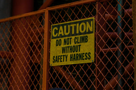 Yellow caution sign on a metal fence reading 'Do Not Climb Without Safety Harness' — emphasizing workplace safety protocols.