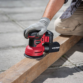 A person wearing a gray work glove uses a red and black Worcraft cordless orbital sander to smooth a wooden beam outdoors on concrete pavement.